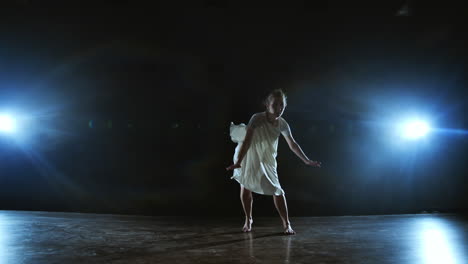 A-young-female-ballerina-barefoot-jumps-on-stage-and-moves-in-slow-motion-in-a-loose-white-dress.